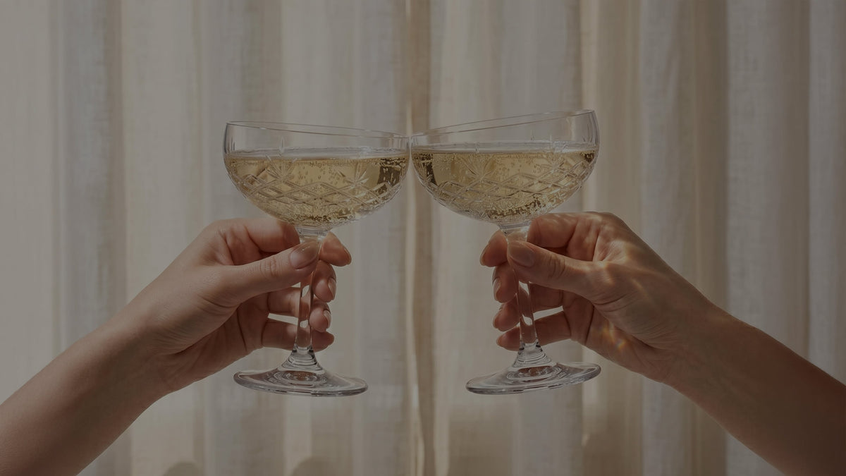 Hands holding two coupe glasses filled with sparkling wine against sheer curtains