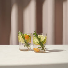Two clear Scandinavian-style water glasses with lemon, herb, and orange slices on a light surface and soft background.