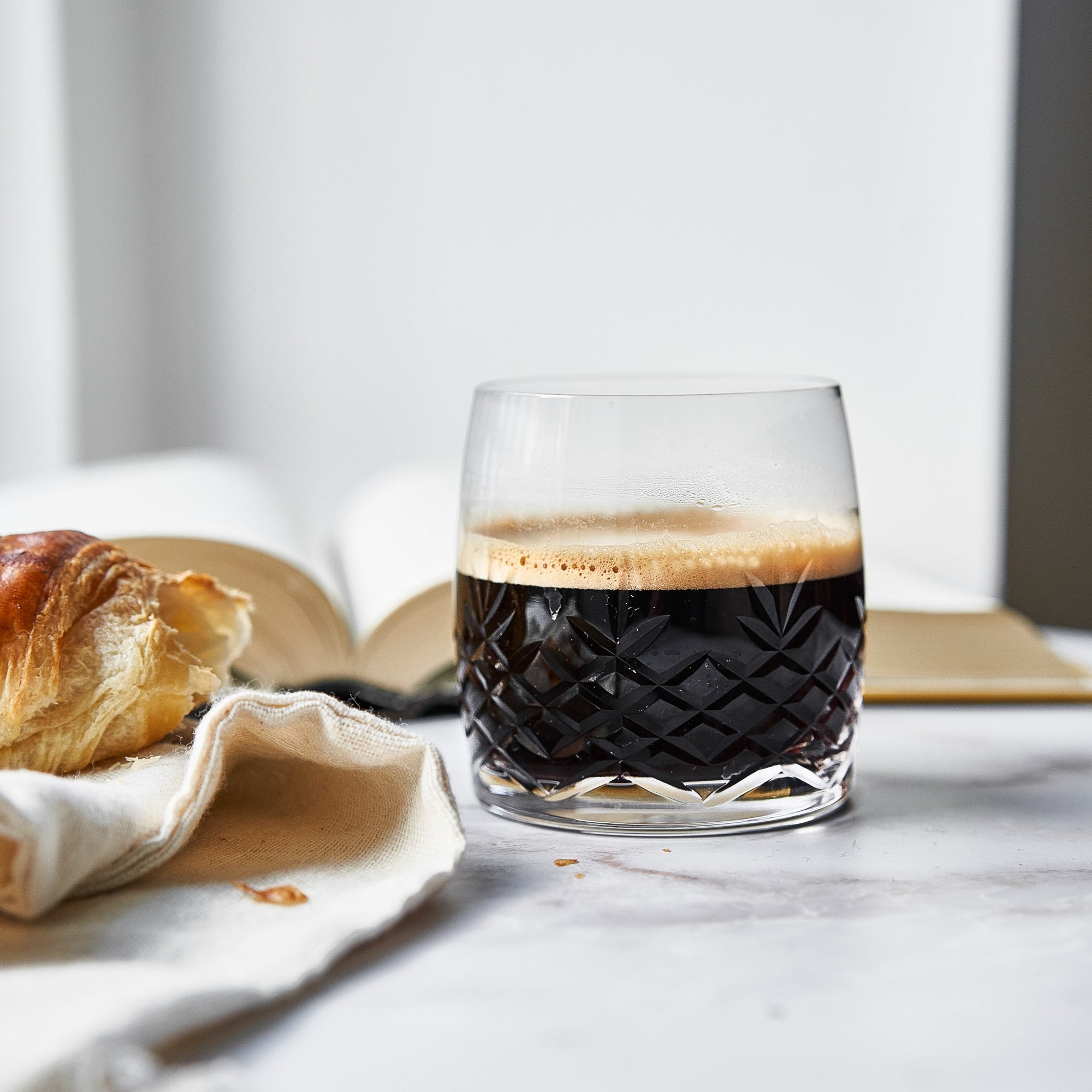 Clear crystal glass with faceted design holding black coffee beside a croissant on a marble surface with open books in background