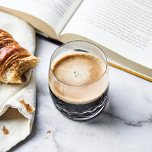 Faceted clear crystal glass with espresso, beside a croissant on a linen napkin and an open book on marble surface
